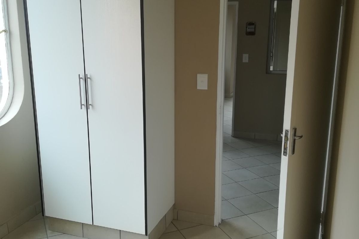 A bright, empty bedroom featuring tiled flooring, a built-in white wardrobe, and an arched window, leading into an adjacent hallway.
