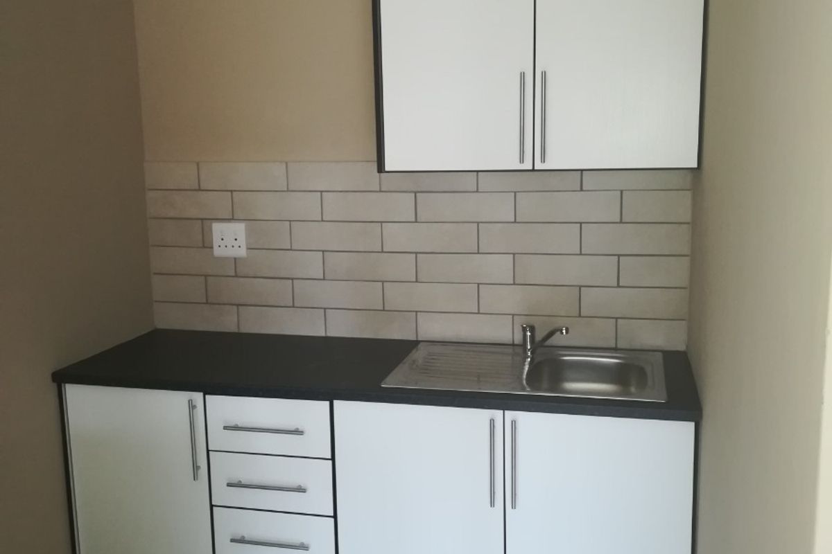 A compact kitchen area featuring white built-in cabinetry, a stainless steel sink, subway tile backsplash, and neutral tiled flooring.
