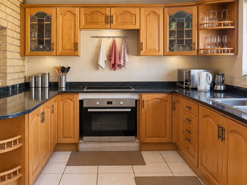 A functional kitchen featuring wooden cabinetry, dark laminate countertops, a built-in oven, electric cooktop, and a brick feature wall.