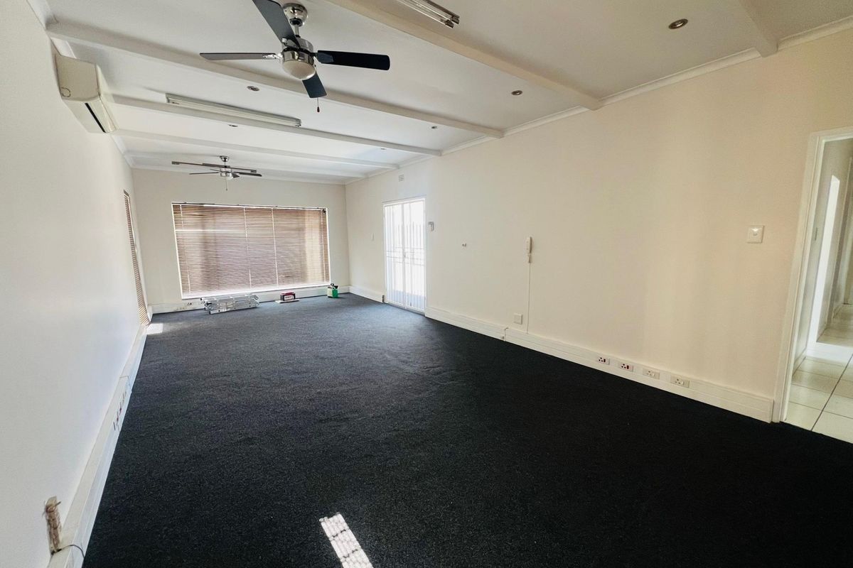 Spacious, empty commercial living area featuring dark carpeting, ceiling fans, air conditioning, and large windows with venetian blinds.