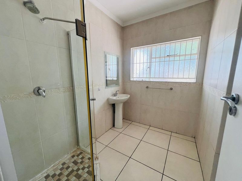 A functional bathroom featuring a pedestal basin, a glass-enclosed shower, and neutral wall and floor tiling with security bars on the window.