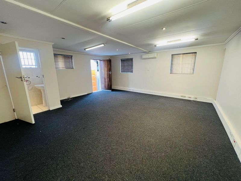 A spacious, empty commercial office area featuring dark carpeting, fluorescent lighting, a wall-mounted air conditioner, and an attached private washroom with a pedestal sink.