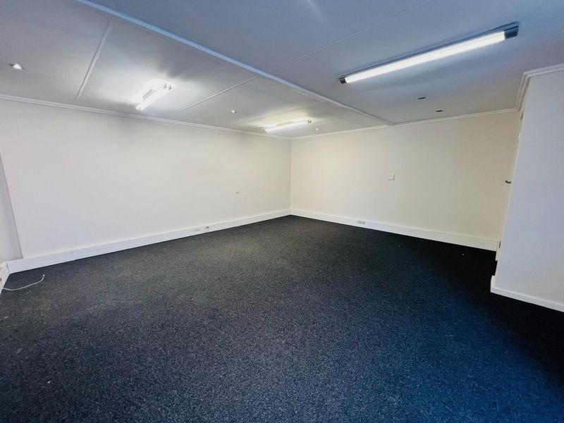 A clean, empty commercial office space featuring neutral walls, dark carpeting, and functional overhead fluorescent lighting.