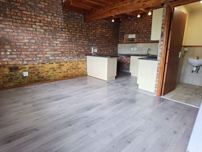 Open plan living area featuring exposed brick walls, timber ceiling beams, and a compact kitchen with laminate flooring throughout.