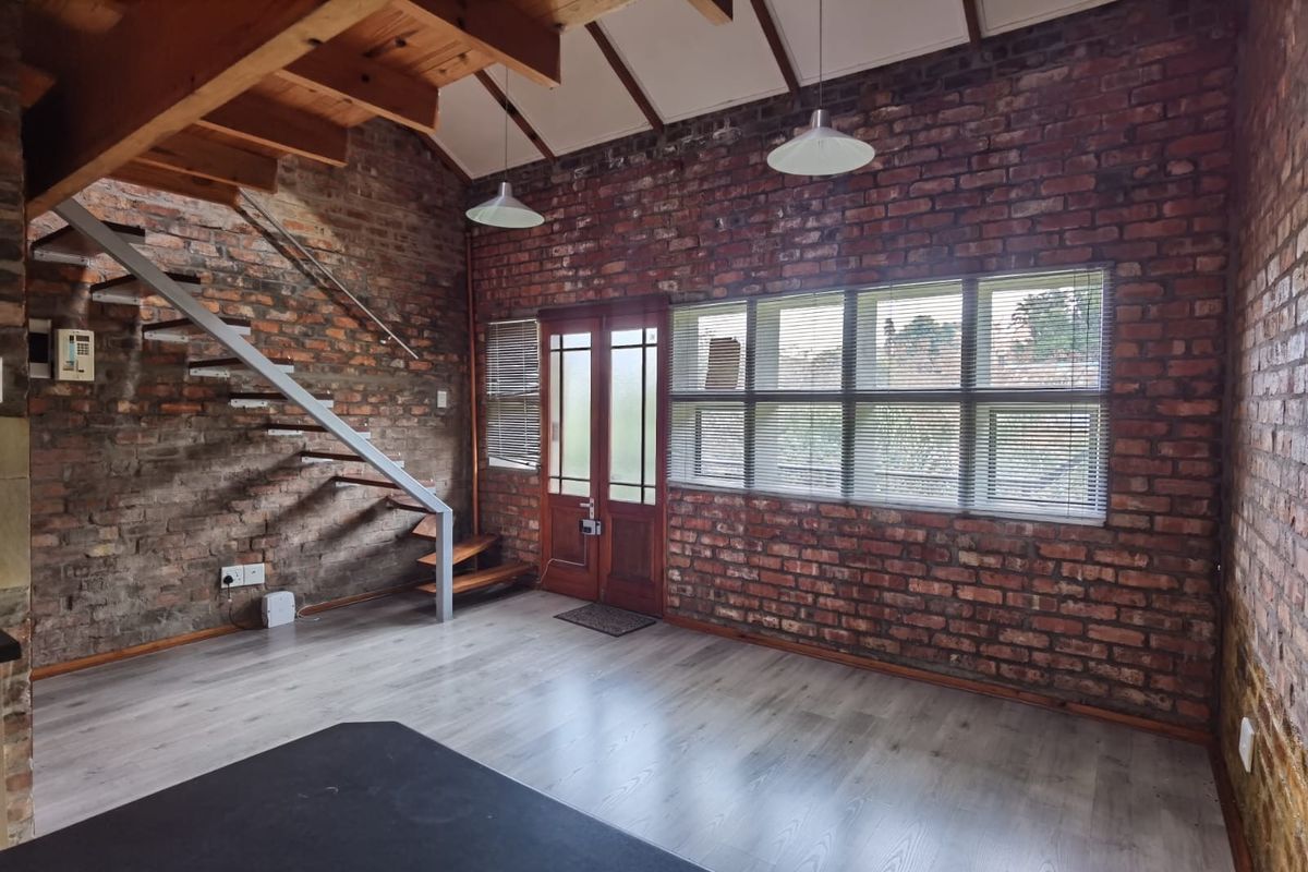 Spacious living area featuring exposed brick walls, timber staircase, and laminate flooring, offering a rustic industrial aesthetic with abundant natural light.