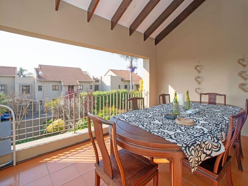 A covered balcony featuring exposed timber rafters, tiled flooring, and a metal balustrade, offering an open-air dining space with views of the surrounding complex.