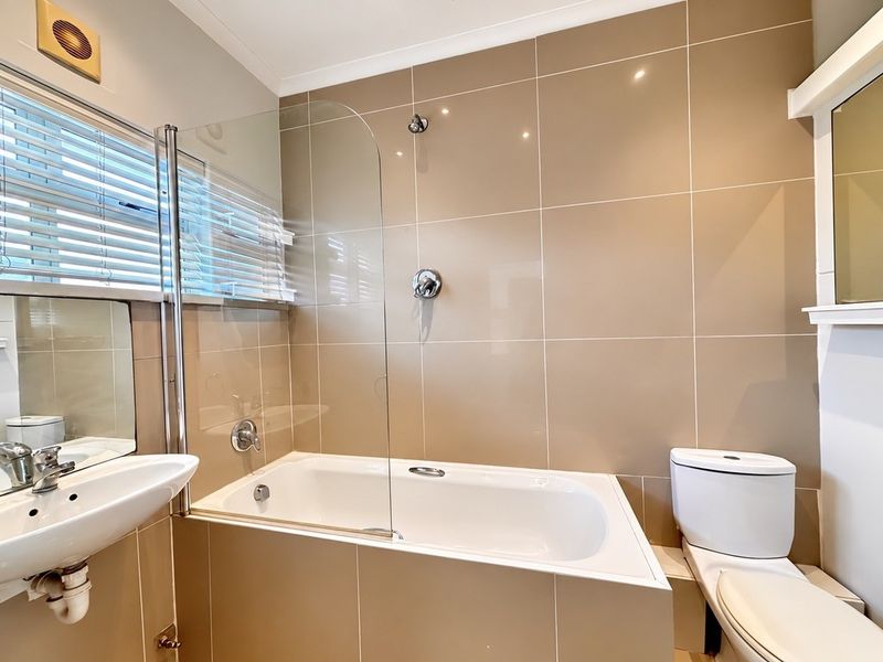 A clean, modern bathroom featuring a built-in bathtub with a glass shower screen, pedestal basin, and toilet, finished with neutral wall tiles.
