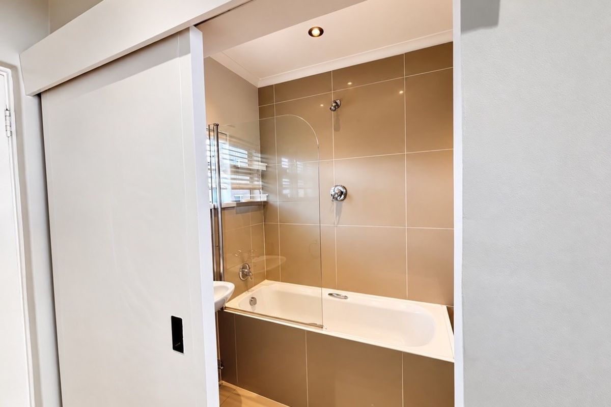 A modern bathroom featuring a built-in bathtub with a glass shower screen, neutral wall tiling, and recessed lighting, accessed via a sliding door.