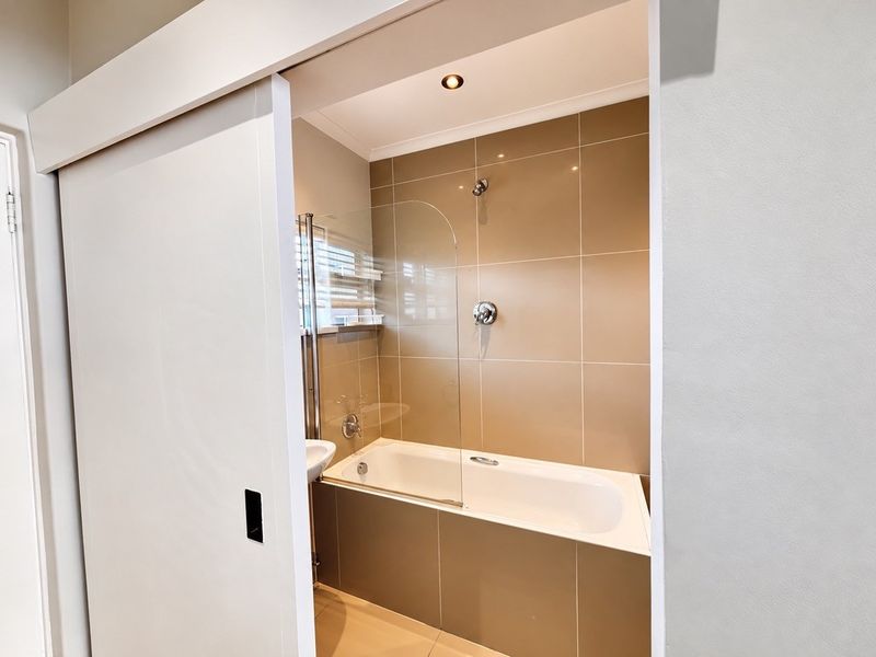 A modern bathroom featuring a built-in bathtub with a glass shower screen, neutral wall tiling, and recessed lighting, accessed via a sliding door.