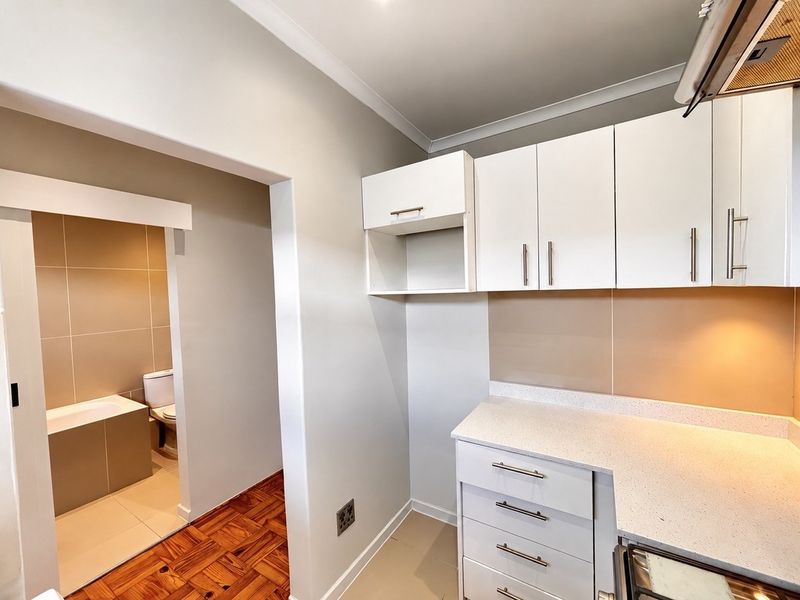 A clean, modern kitchen area featuring white cabinetry, stone countertops, and an extractor fan, with an adjacent bathroom visible through the doorway.