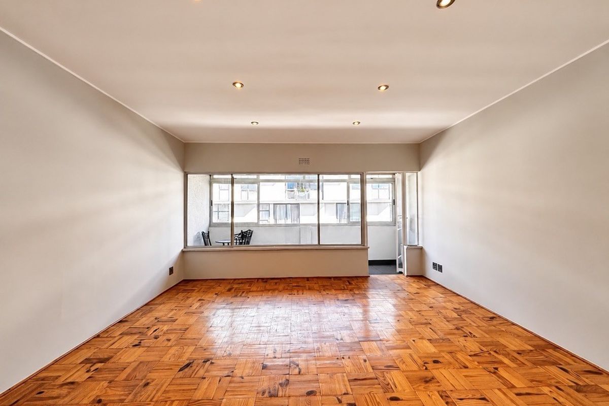 A bright, empty living room featuring classic parquet flooring, recessed downlighting, and large windows providing natural light and access to an adjoining balcony.