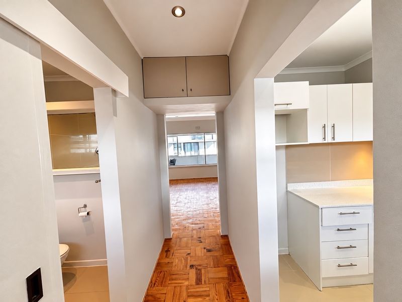 A bright hallway featuring parquet flooring, connecting to a modern kitchen with white cabinetry and stone countertops, leading towards an open living area.