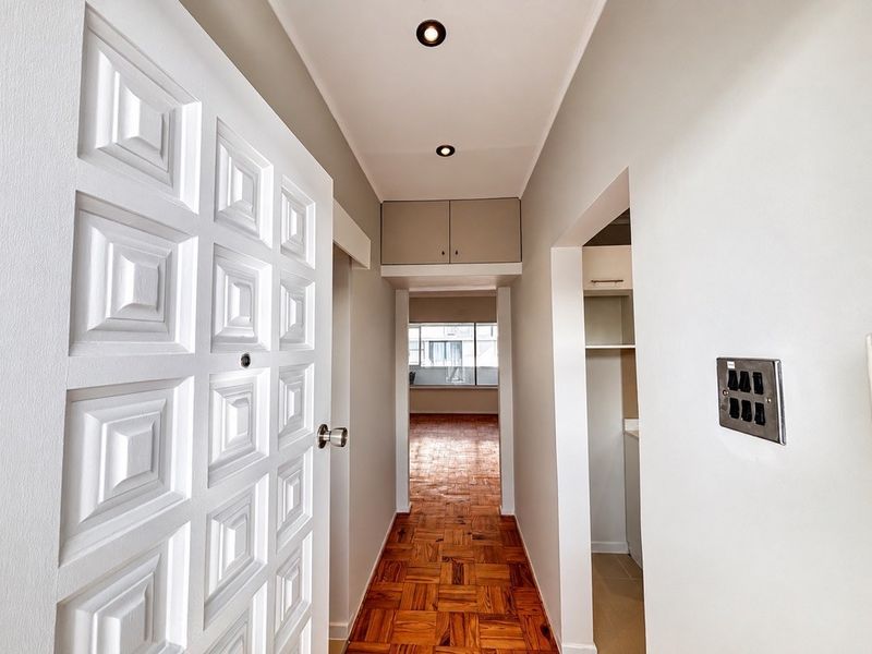 A bright, clean hallway featuring classic parquet flooring, recessed downlighting, and a built-in overhead storage cupboard, leading towards an open living area.