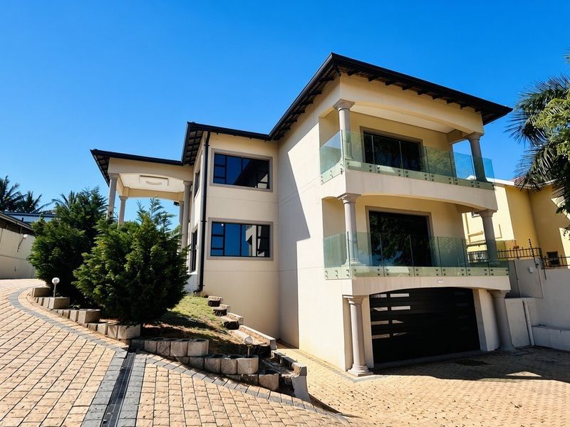 A modern double-storey residential property featuring a paved driveway, glass-railed balconies, and a secure garage entrance.