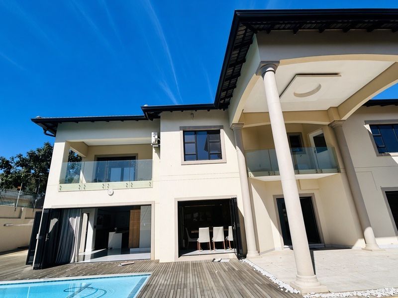 Rear elevation of a modern double-story home featuring a swimming pool, timber decking, covered patio, and glass balustrades with large bi-fold doors.