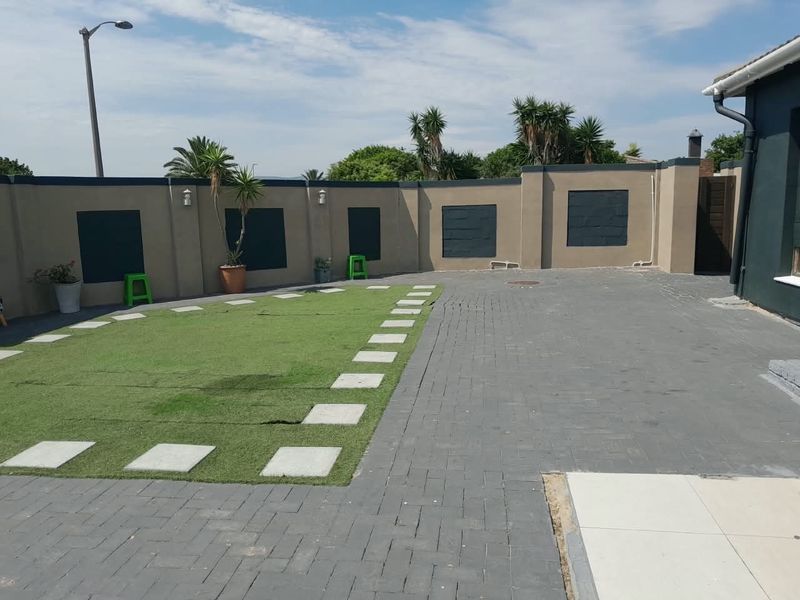 A rear courtyard area featuring a paved driveway, artificial lawn with stone pavers, and a secure perimeter wall with integrated lighting.
