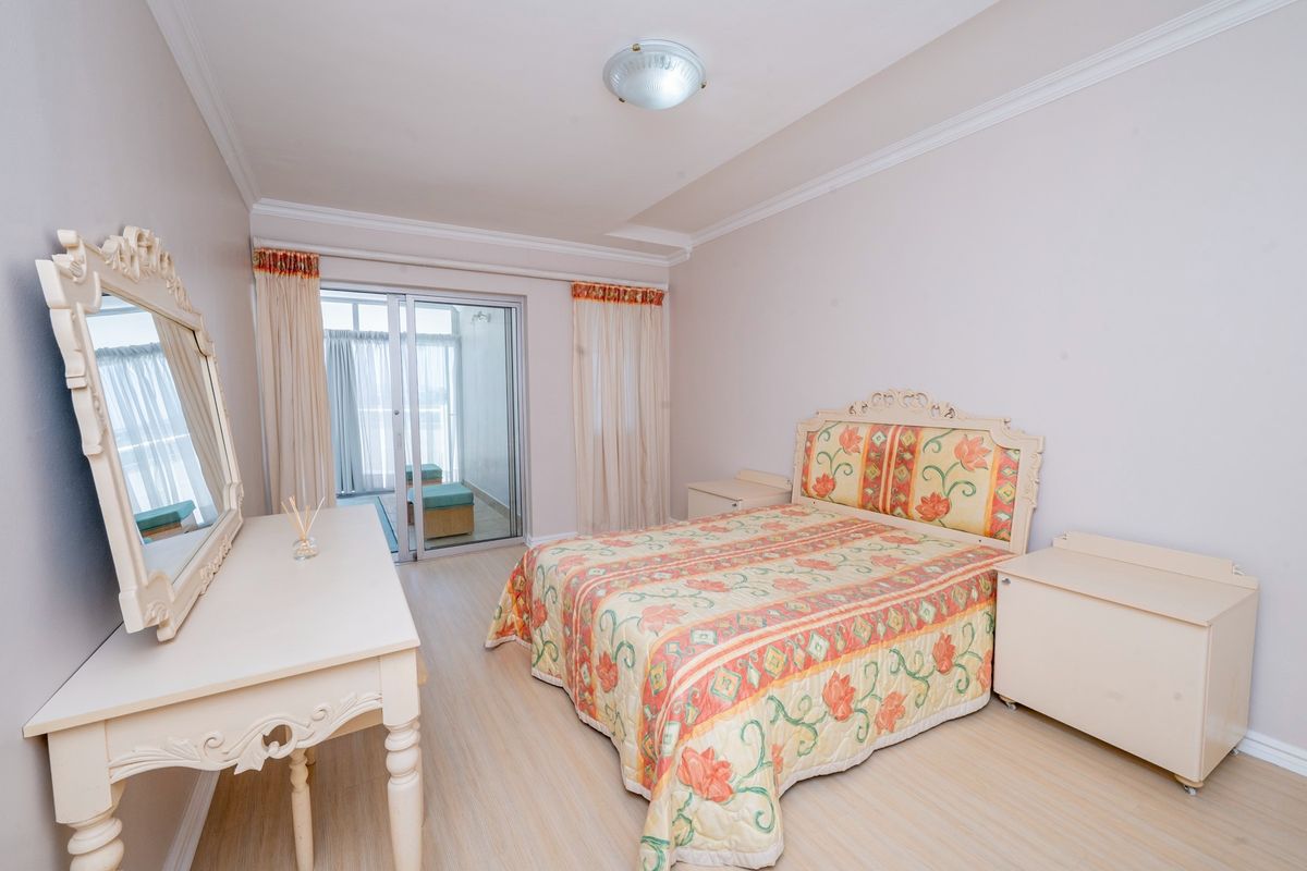 A well-lit bedroom featuring light-toned laminate flooring, ceiling cornicing, and a sliding glass door providing access to an attached balcony.