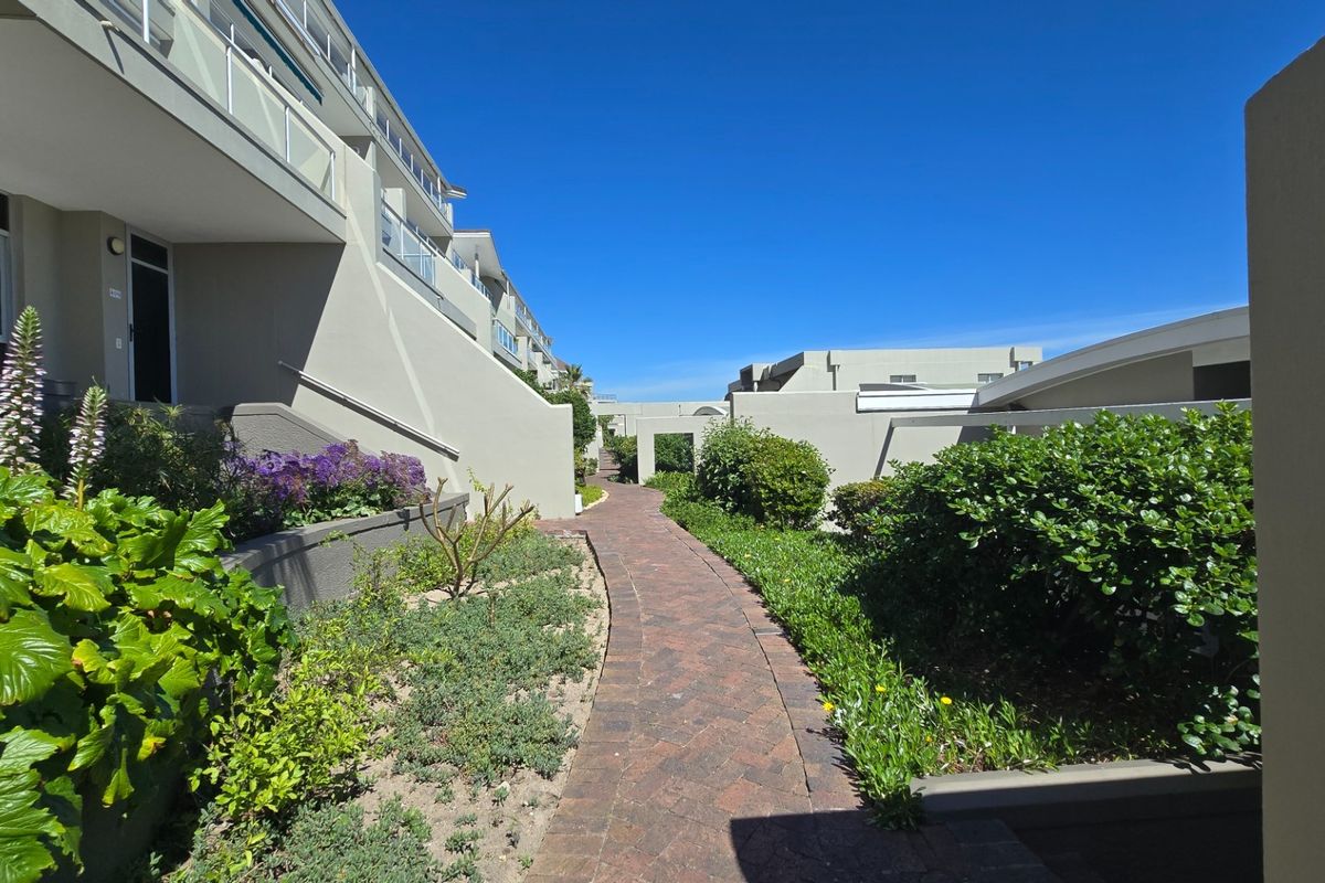 A well-maintained exterior pathway leading through a landscaped garden area between apartment building blocks, featuring paved walkways and established garden beds.