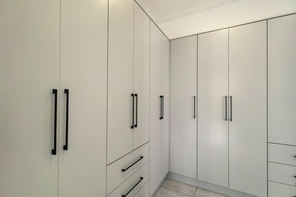 A clean, modern bedroom featuring extensive floor-to-ceiling built-in cupboards with contemporary black hardware and neutral tiled flooring.