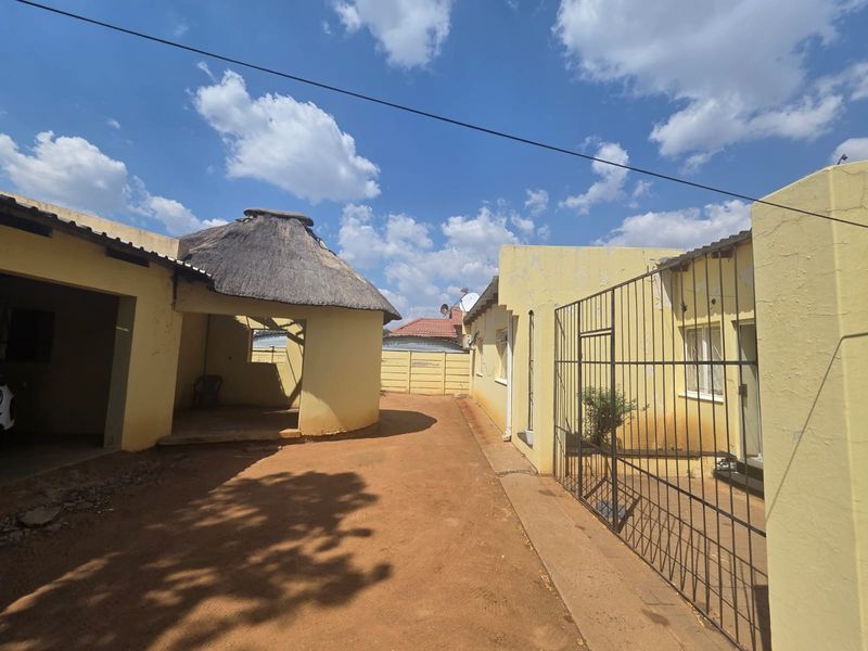 Rear exterior view showing a detached thatch lapa, concrete driveway, and perimeter walling with visible wear to the exterior paintwork.