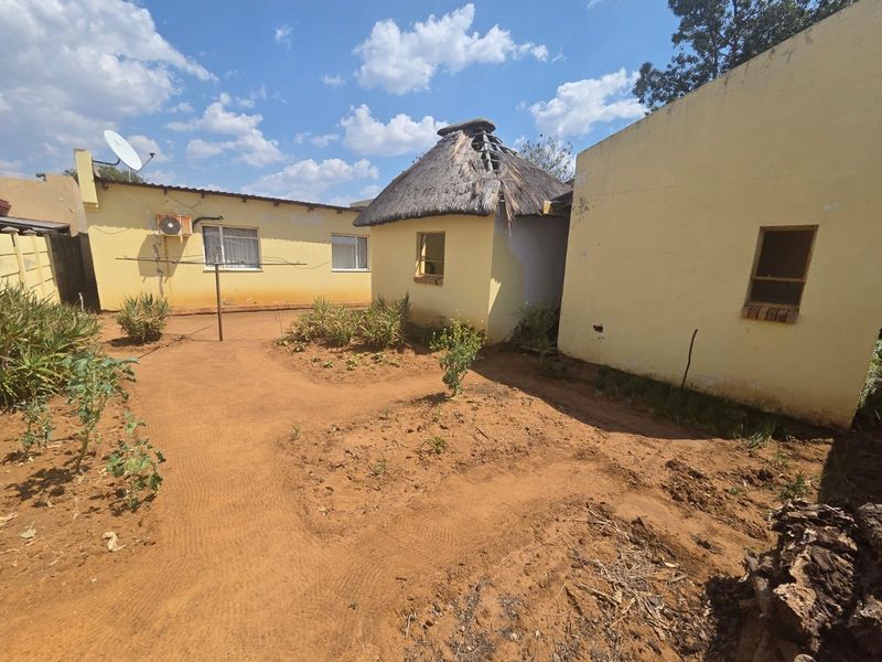 Rear exterior view of the property showing a garden area, plastered masonry walls, and a thatch-roofed structure requiring significant maintenance and repair.