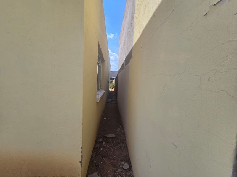 A narrow side passage between two exterior walls showing signs of plaster cracking, paint deterioration, and unpaved ground.