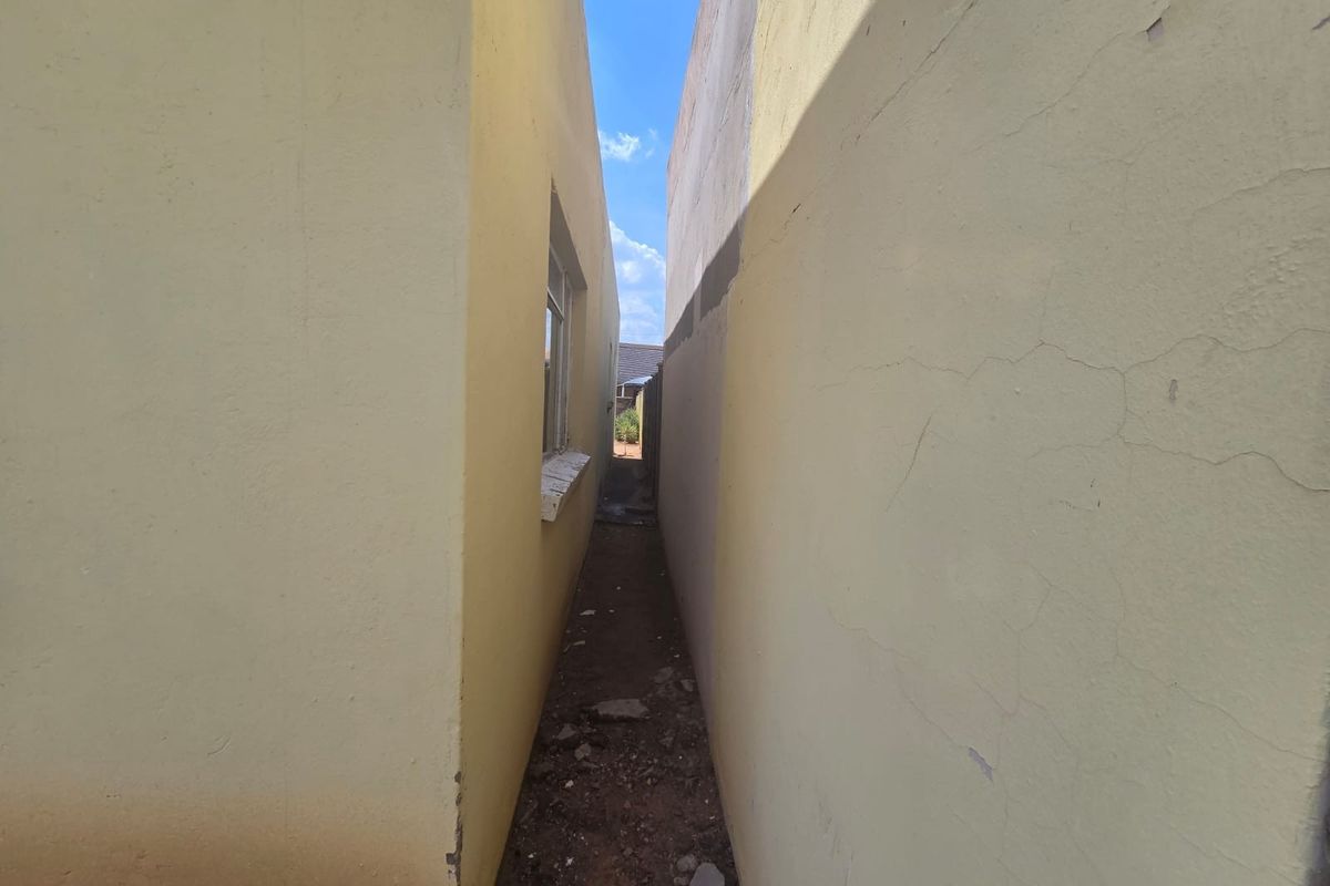 A narrow side passage between two exterior walls showing signs of plaster cracking, paint deterioration, and unpaved ground.