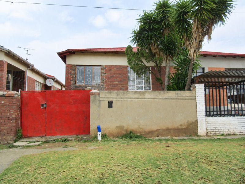 Front elevation of a brick house featuring a red metal roof, security gate, and boundary wall with established garden elements.
