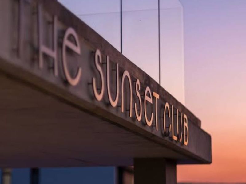 A close-up view of the modern concrete signage for The Sunset Club, featuring a sleek glass balustrade against a sunset sky.