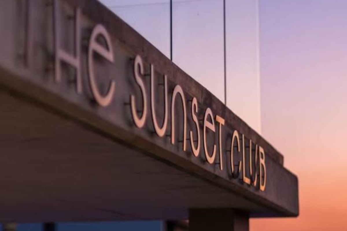 A close-up view of the modern concrete signage for The Sunset Club, featuring a sleek glass balustrade against a sunset sky.
