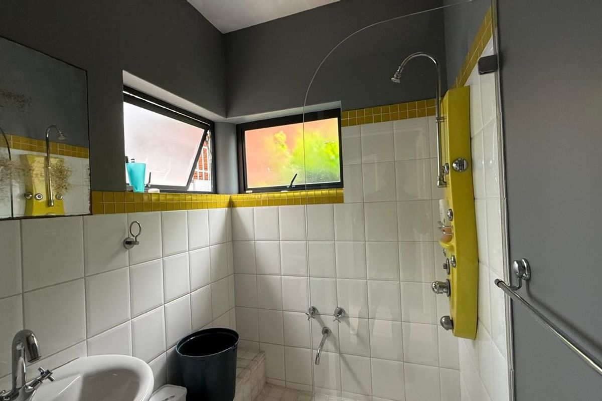 A functional bathroom featuring a tiled shower enclosure with a wall-mounted panel, pedestal basin, and casement windows providing natural light.