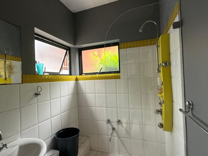 A functional bathroom featuring a tiled shower enclosure with a wall-mounted panel, pedestal basin, and casement windows providing natural light.