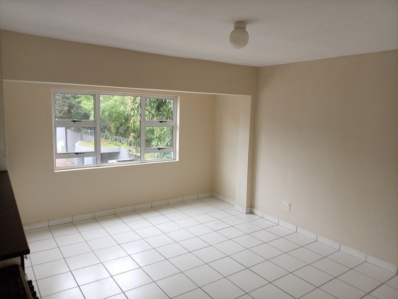 A bright, empty living area featuring neutral walls, white tiled flooring, and large aluminium-framed windows providing natural light.