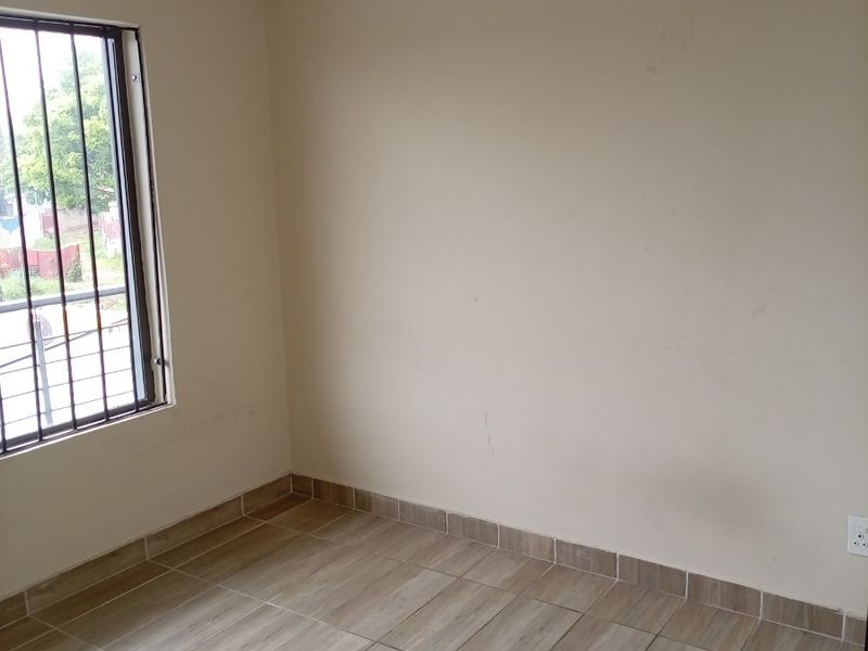 A clean, empty bedroom featuring neutral tiled flooring, security bars on the window, and plain walls, ready for personalization.