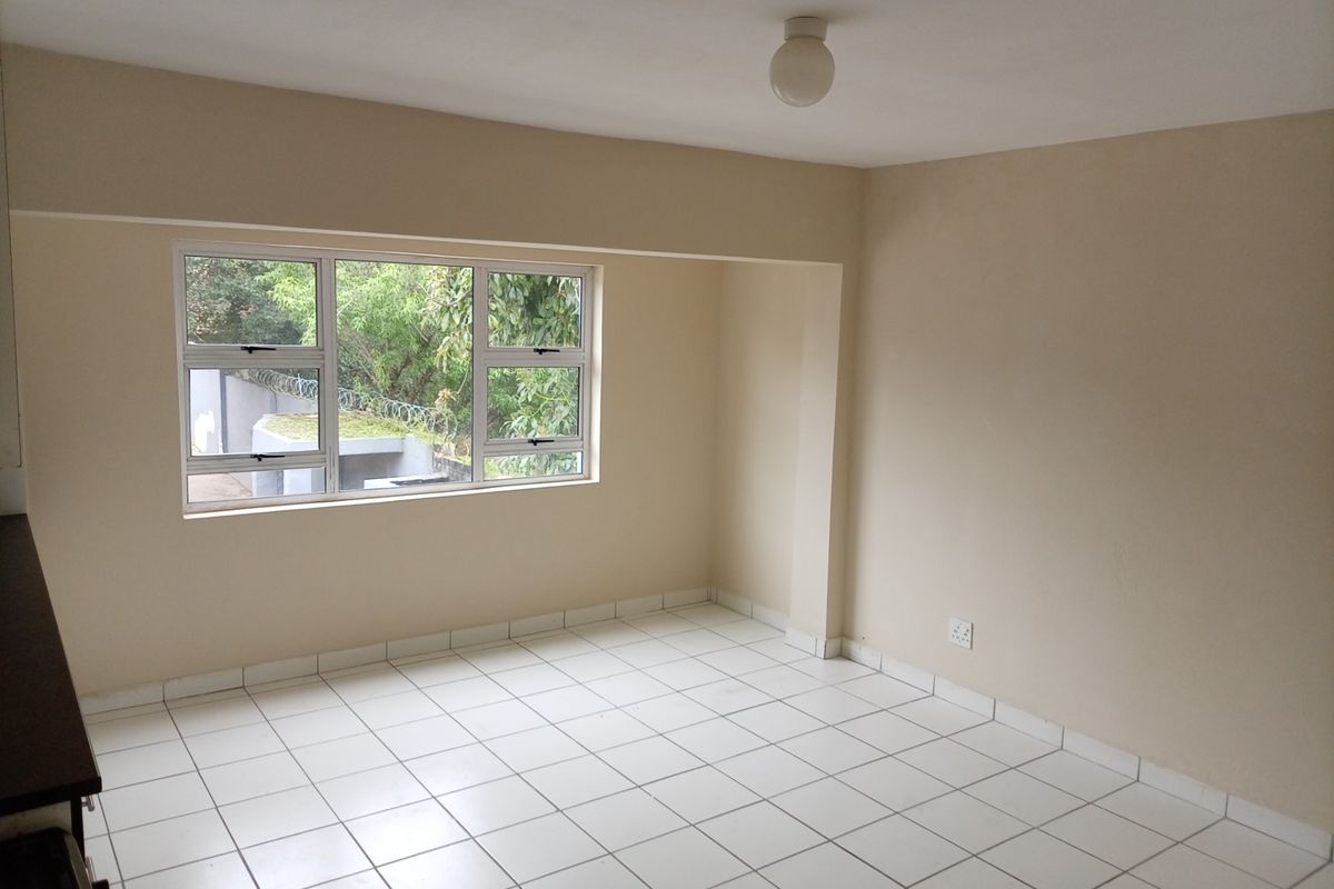 A bright, empty living area featuring neutral walls, white tiled flooring, and large aluminium-framed windows providing natural light.