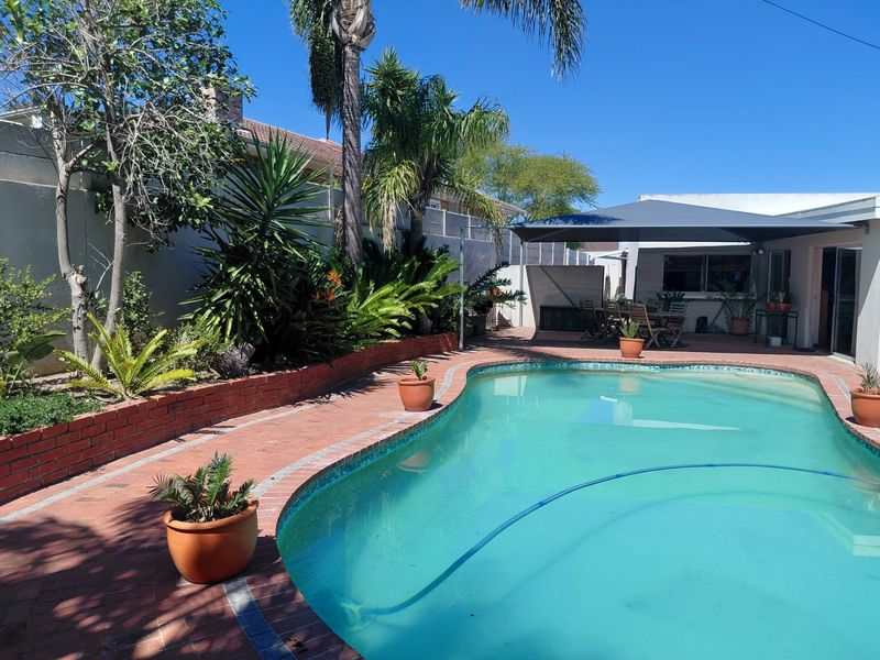 Outdoor pool area featuring a swimming pool, brick paved surround, and a covered patio space, set within a landscaped garden with boundary walls.