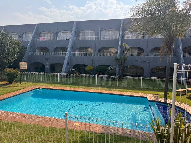 A communal swimming pool area featuring a brick-edged pool, metal fencing, and a view of the multi-story apartment building facade with balconies.