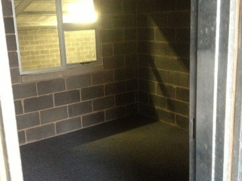 A small, empty office space featuring exposed concrete block walls, carpeted flooring, and a suspended ceiling, suitable for a workspace or storage area.
