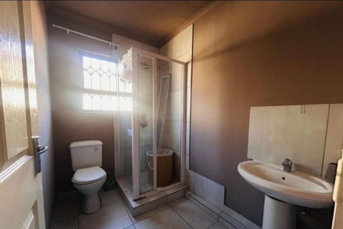 A compact bathroom featuring a shower enclosure, pedestal basin, and toilet, with tiled flooring and security bars on the window.