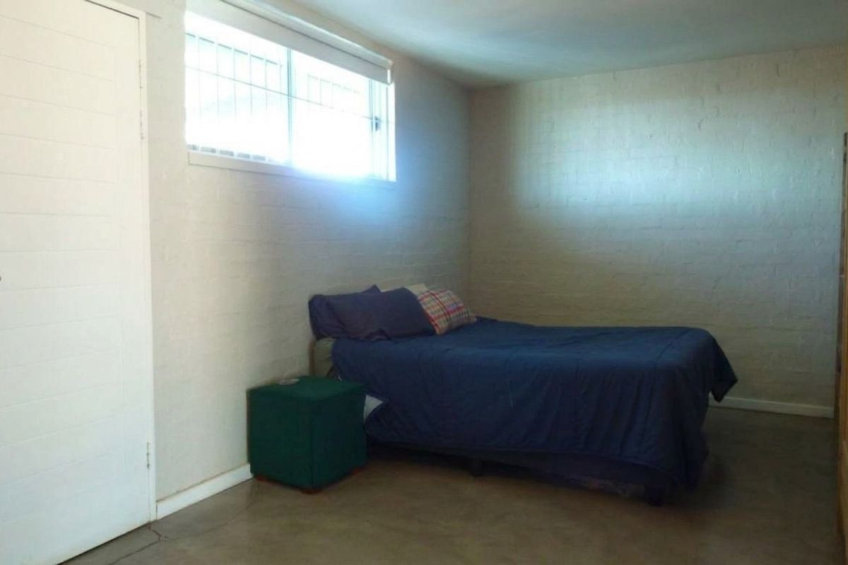 A simple bedroom featuring painted brick walls, concrete flooring, and a window fitted with security bars.