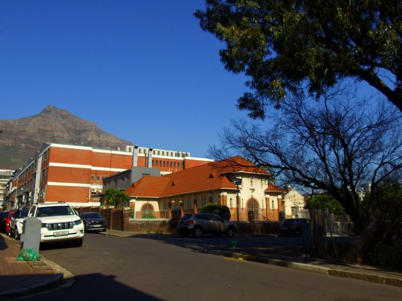 A street-level view of the neighbourhood approach, featuring paved sidewalks, asphalt road, and surrounding brick boundary walls with mountain views in the background.