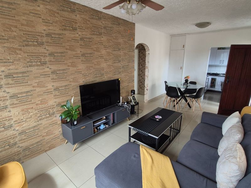 Open-plan living area featuring a textured stone-cladding feature wall, tiled flooring, and a ceiling fan, with an adjacent dining space and kitchen access.
