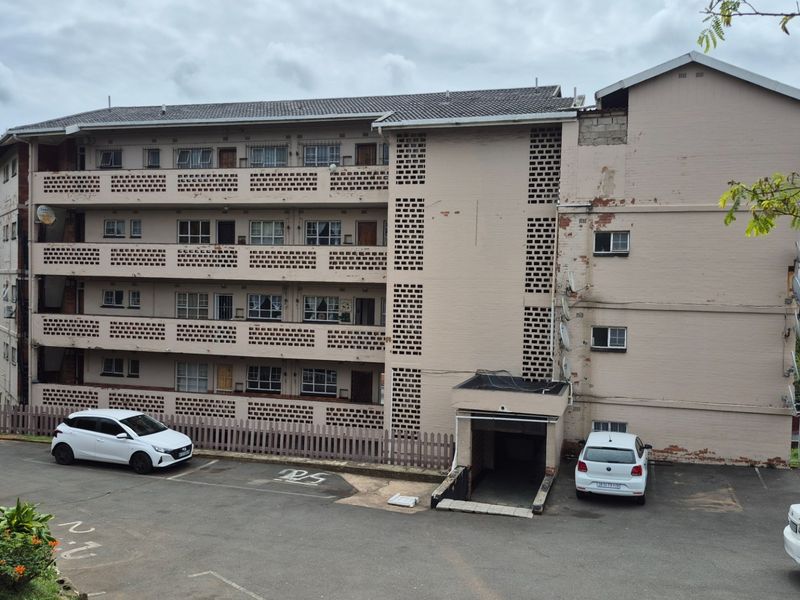 Exterior view of a multi-story apartment building featuring concrete breeze block balustrades and a paved parking area.