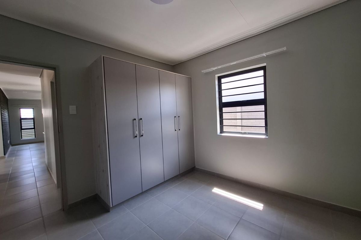 A clean, empty bedroom featuring neutral tiled flooring, built-in cupboards, and an aluminium-framed window, offering a bright and versatile living space.