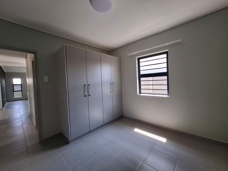 A clean, empty bedroom featuring neutral tiled flooring, built-in cupboards, and an aluminium-framed window, offering a bright and versatile living space.