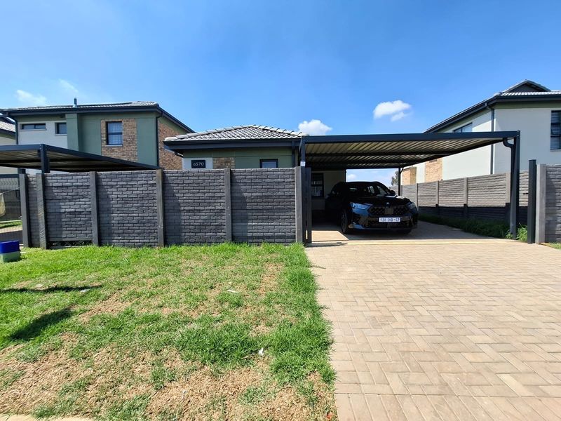 Front view of the townhouse featuring a paved driveway, a modern carport structure, and a secure brick boundary wall.