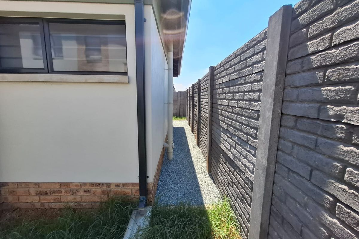 A side passage of the townhouse featuring a gravel pathway, a precast concrete boundary wall, and a functional downpipe system.