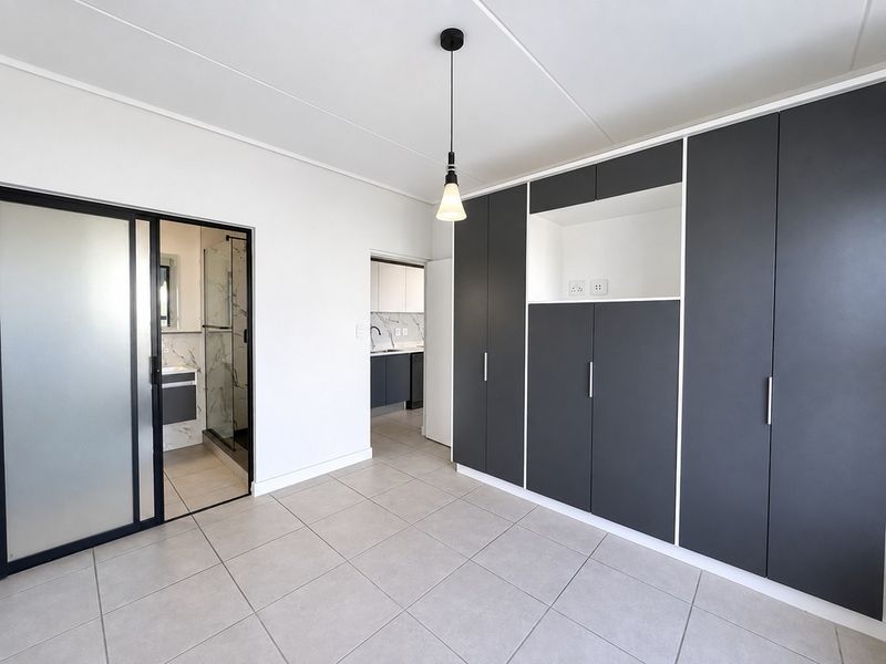 A modern, empty bedroom featuring sleek dark built-in cupboards, neutral tiled flooring, and direct access to an en-suite bathroom.
