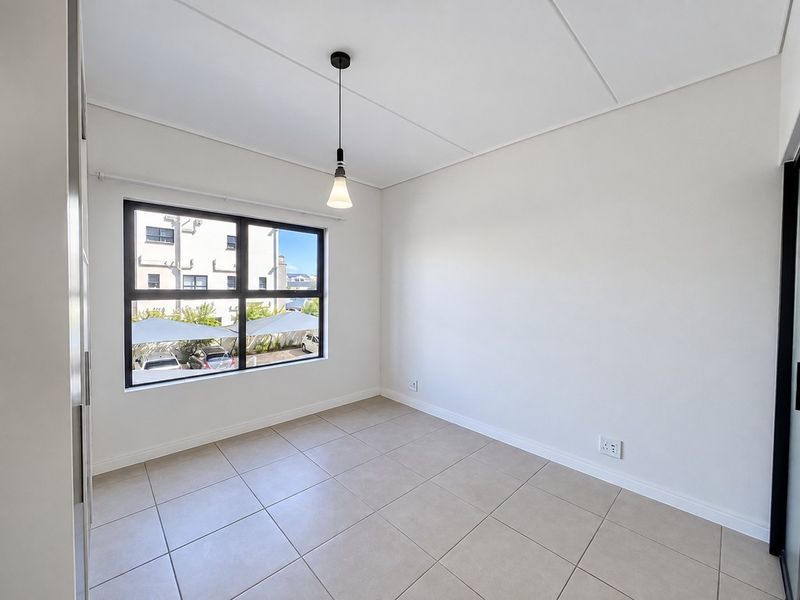 A bright, empty bedroom featuring neutral tiled flooring, a modern pendant light, and a large window with black frames.