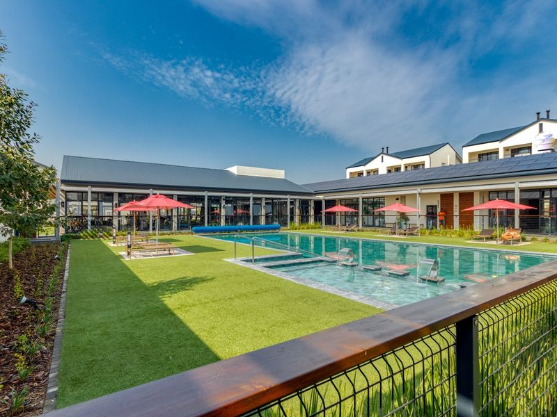 A well-maintained communal swimming pool area featuring a modern pool with water features, artificial lawn, and surrounding building structures with glass facades.